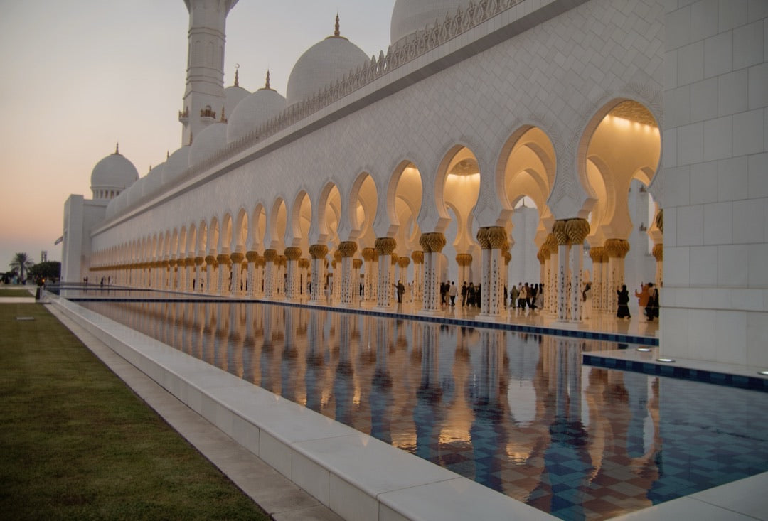 Arched colonnade of a white grand mosque reflected in a pool at dusk